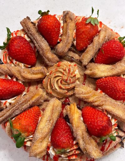 A beautifully decorated dessert features a circular arrangement of fresh strawberries, whipped cream rosettes, and sugar-coated churros atop a layer of drizzled red sauce. The contrast of colors and textures creates an appealing and appetizing presentation.