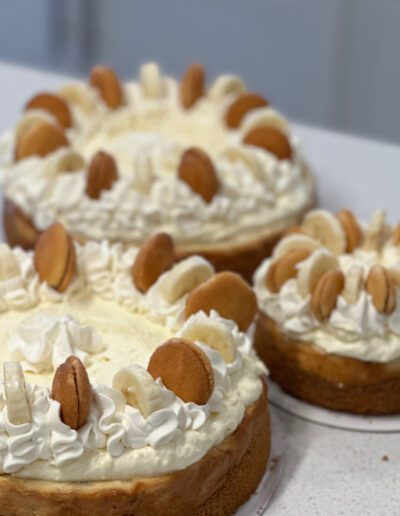 Three cheesecakes topped with whipped cream rosettes, vanilla wafers, and banana slices are displayed on a white countertop. The cheesecakes vary in size and have a graham cracker crust. The background shows white cabinets and a kitchen setting.