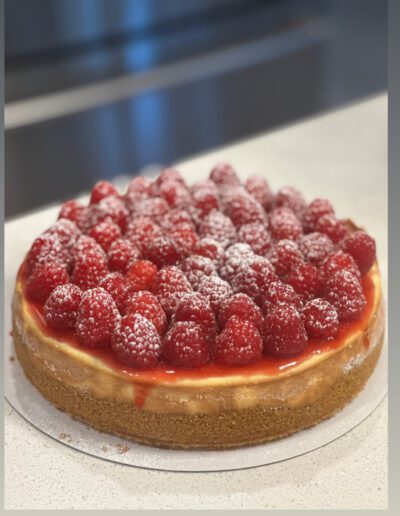 A cheesecake with a golden brown crust topped with a generous layer of fresh raspberries. The raspberries are sprinkled with powdered sugar, adding a touch of sweetness. The cake sits on a white surface with a blurred background.