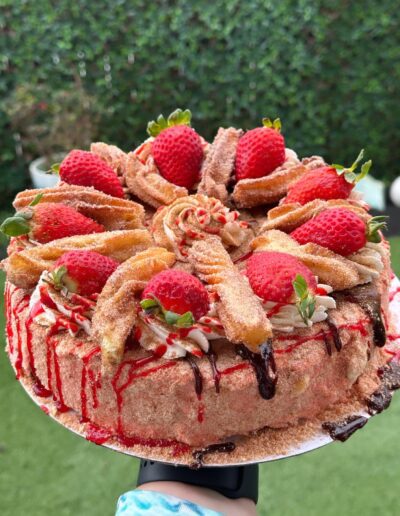 A beautifully decorated cake is topped with fresh strawberries, cinnamon sugar-coated churros, swirls of cream, and drizzles of red and chocolate syrup. The background shows a vibrant green garden.