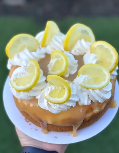 A close-up of a lemon cheesecake topped with swirls of whipped cream and lemon slices. The cake is drizzled with a golden syrup and is held on a white plate against a blurred green background.