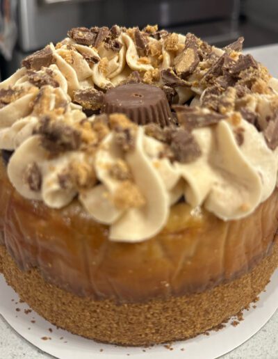 A cheesecake topped with a caramel layer, swirls of whipped cream, and large chunks of chocolate and peanut butter cups, resting on a graham cracker crust. The cake is placed on a white, round cake board with a kitchen blurred in the background.