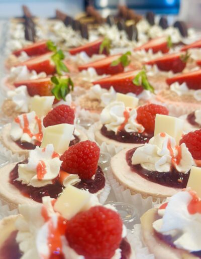 A close-up of various desserts arranged on a platter. The foreground features mini cheesecakes topped with whipped cream, raspberry sauce, fresh raspberries, and white chocolate. Background desserts include chocolate-covered pieces and treats garnished with strawberries.