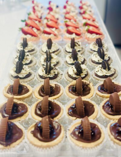 A display of assorted mini desserts arranged in rows. The front row has chocolate tarts with chocolate filling and a piece of chocolate candy on top. The middle row features vanilla cupcakes topped with whipped cream and an Oreo cookie. The back row holds strawberry-topped cupcakes.