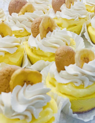 A close-up of lemon cupcakes topped with swirls of white frosting, a crispy wafer, and a banana chip. The cupcakes are arranged in rows and have a vibrant yellow color, giving them a fresh and appetizing appearance.