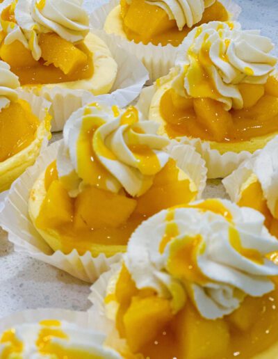 Close-up of several mini cheesecakes topped with dollops of whipped cream, mango slices, and a drizzle of mango syrup. Each cheesecake sits in a white fluted paper cup, arranged closely together on a white surface.