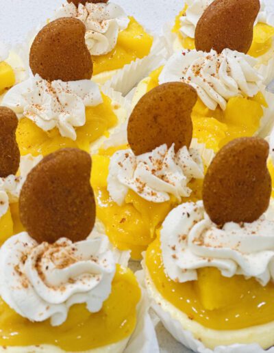 Close-up of several dessert cups filled with creamy yellow pudding, topped with whipped cream, a dusting of cinnamon, and a round cookie. Each dessert is presented in a white paper cup. The appearance is inviting and festive.