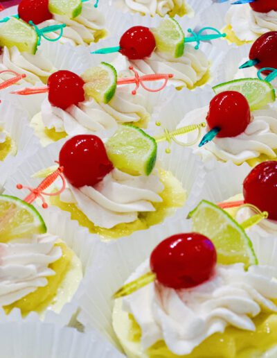 A close-up of cupcakes decorated with white frosting, red cherries, and lime wedges. The cupcakes are arranged in rows, each topped with a colorful, translucent plastic cocktail pick. The background shows more cupcakes, creating a visually appealing display.