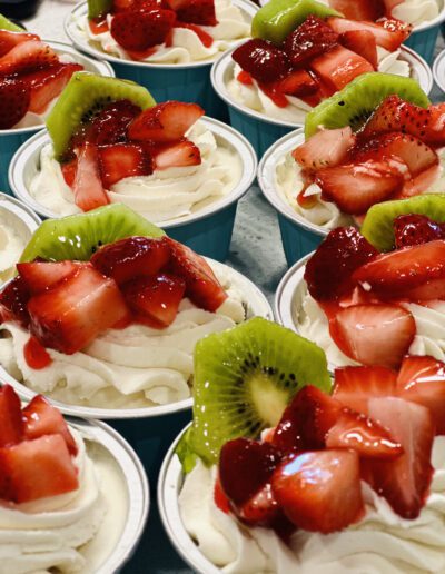 A close-up of several dessert cups, each filled with whipped cream, topped with diced strawberries and kiwi slices. The fruits glisten with a light syrup, making the vibrant reds and greens stand out against the creamy white backdrop. The cups are blue.