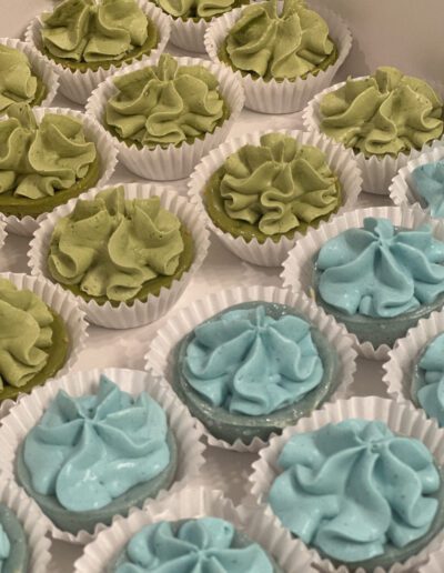 A display of small cupcakes with swirls of green and blue frosting, each placed in white paper liners. The cupcakes are arranged closely together on a flat surface.