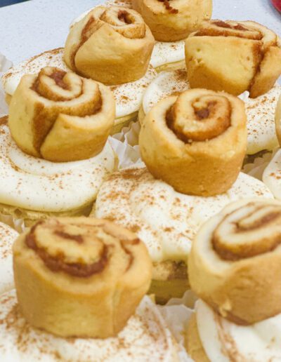 A close-up of several cinnamon rolls. Each roll is placed on a layer of white icing and sprinkled lightly with cinnamon. The golden-brown swirls are soft and inviting, suggesting a freshly baked, sweet aroma. They are arranged neatly on a white surface.