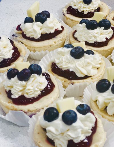 A close-up of a batch of mini cheesecakes topped with whipped cream, blueberry compote, fresh blueberries, and a small piece of white chocolate. They are arranged on a white surface in individual white paper wrappers.