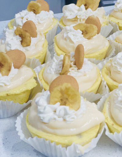 Cupcakes topped with swirls of creamy frosting, banana slices, and vanilla wafer cookies are displayed in white paper liners. The frosting looks smooth and thick, contrasting with the crunchy toppings. The cupcakes have a light yellow hue, suggesting a banana flavor.