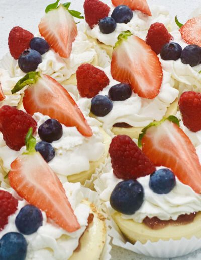 Mini cheesecakes topped with generous swirls of whipped cream, fresh strawberries, raspberries, and blueberries, arranged on a white surface. Each cheesecake is nestled in a white paper liner, creating a vibrant and appetizing display of desserts.