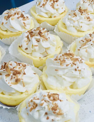 A close-up image of several lemon cheesecake cupcakes topped with whipped cream and toasted coconut flakes. The cupcakes are arranged in white cupcake liners on a light-colored surface. The desserts have a creamy texture and look freshly made.