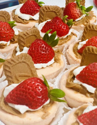 Mini cheesecakes topped with fresh strawberries, a dollop of whipped cream, and a cookie piece. The cheesecakes are arranged in white paper cups, showcasing their detailed toppings and inviting presentation.