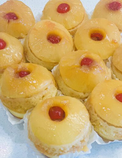 A close-up view of several pineapple upside-down mini cakes. Each cake has a pineapple ring with a maraschino cherry in the center, placed on top of golden, caramel-glazed sponge cakes. The cakes are individually placed in white paper cups.