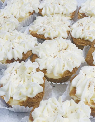 A close-up of multiple cupcakes topped with swirls of creamy white frosting. The cupcakes are placed in white paper liners and appear to be dusted lightly with a fine powder, possibly nutmeg or cinnamon. The frosting is piped decoratively on each cupcake.