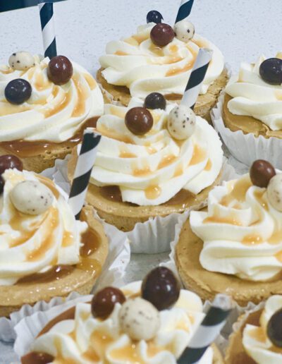 A close-up of several cupcakes decorated with swirls of white frosting, drizzles of caramel sauce, various chocolate-coated spherical candies, and black-and-white striped paper straws. The cupcakes are arranged side by side on a white surface.