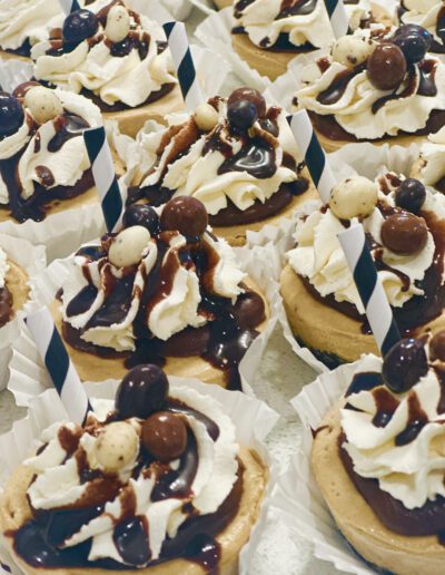 A display of cupcakes topped with swirls of whipped cream, chocolate drizzle, and various spherical chocolate candies. Each cupcake has a black and white striped straw inserted into the frosting, and they are arranged in white paper liners.