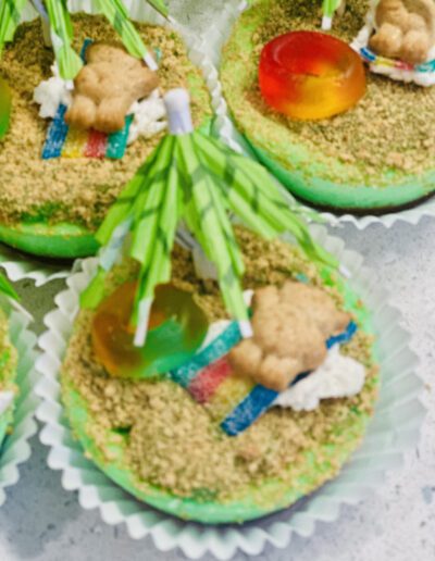 A cupcake designed like a tropical island with green icing, crushed graham crackers as sand, a teddy bear cookie lounging under a green paper umbrella, a gummy ring as a float, and colorful candy strip representing a beach towel.