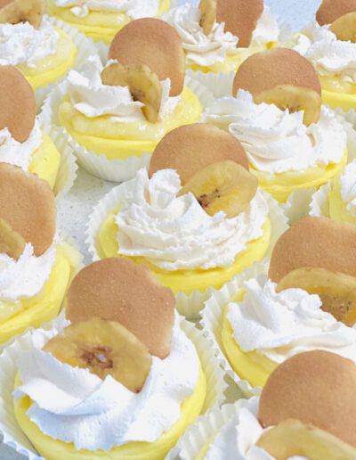 A tray of mini banana pudding desserts in white paper cups. Each dessert features a swirl of whipped cream, a vanilla wafer cookie, and a banana slice garnishing the top. The yellow pudding is visible underneath the toppings.