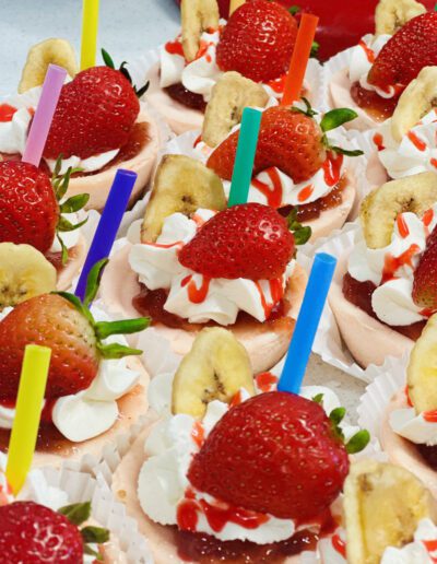 A colorful array of mini desserts arranged on a tray. Each dessert is topped with whipped cream, a fresh strawberry, banana chips, and a drizzle of red sauce. Different colored straws are inserted into each treat, enhancing the festive presentation.