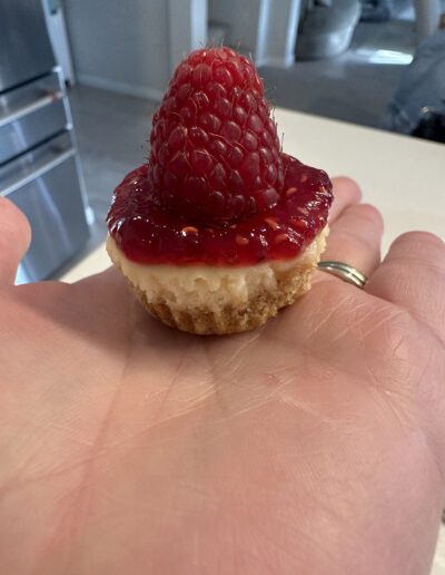 A mini cheesecake topped with a dollop of raspberry sauce and a fresh raspberry sits on the palm of a person's hand. The background shows a modern kitchen with stainless steel appliances and light-colored countertops.