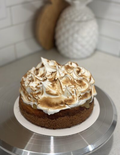 A close-up of a homemade cake on a cake stand. The cake has a browned, swirled meringue topping. In the background, there are two decorative ceramic pineapples on a kitchen countertop with a white tile backsplash.