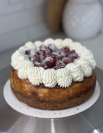 A cheesecake sits on a metal stand. The cake has a golden, caramelized top, a crumbly crust, and is decorated with swirls of whipped cream along the edge. The center is topped with fresh raspberries and a dusting of powdered sugar.