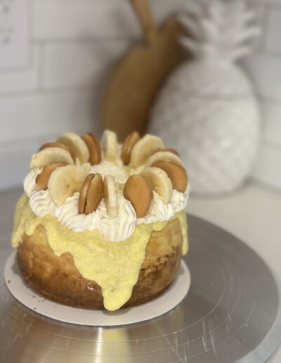 A delicious-looking cake with creamy yellow frosting is decorated with whipped cream, banana slices, and round vanilla wafer cookies on top. The cake sits on a metal platter against a white-tiled background with decor shaped like a pineapple visible in the back.