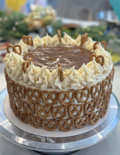 A close-up of a round cake adorned with alternating swirls of cream and mini pretzels on top. The sides are also covered with mini pretzels. The cake appears to have a caramel-like topping in the center and is displayed on a silver cake stand. A blurred background suggests an indoor setting with some greenery.