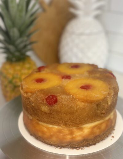 A pineapple upside-down cake sits on a cake stand, decorated with pineapple rings and maraschino cherries. In the background, there are whole pineapples and a white pineapple-shaped decor piece. The scene appears to be set in a kitchen.