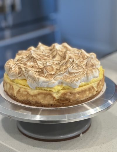 A lemon meringue cheesecake sits on a metal stand. The cheesecake has a golden brown crust, a yellow lemon filling, and is topped with fluffy, browned meringue peaks. The background is softly blurred, focusing attention on the cheesecake.