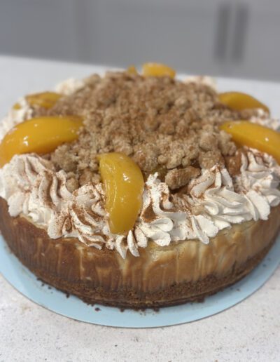 A round peach cobbler cheesecake on a blue plate, topped with whipped cream swirls, peach slices, and a crumbly streusel coating. The cheesecake has a light brown crust and is placed on a white countertop.