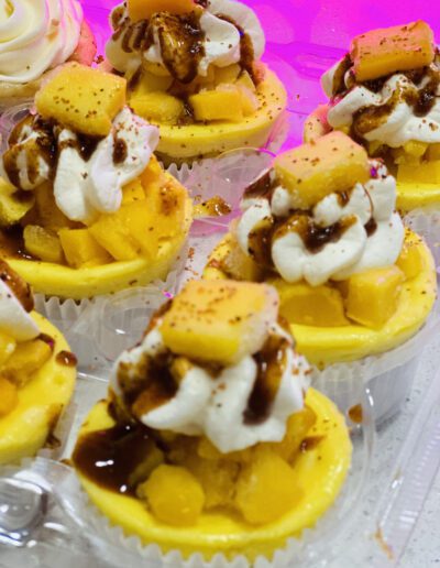 Close-up of several mini cheesecakes topped with whipped cream, chunks of mango, and a drizzle of chocolate sauce. The cheesecakes are arranged in a transparent container, and the background is illuminated with pink light.