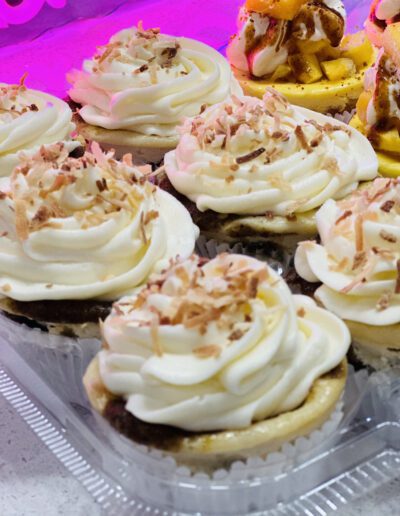 A close-up view of a tray filled with a variety of cupcakes. The cupcakes are topped with swirls of white frosting and sprinkled with what appears to be shredded coconut and chocolate flakes. The background is illuminated with a soft pink light.