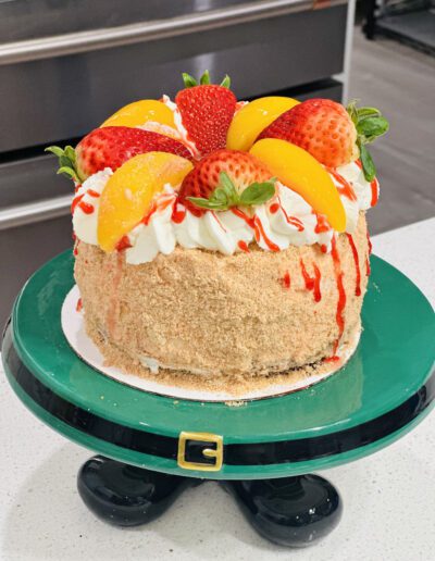 A round cake on a green cake stand with a black belt design. The cake is topped with whipped cream, strawberries, and peach slices, drizzled with red syrup. The sides are coated in crumbs. Background shows a kitchen with a stainless steel oven.