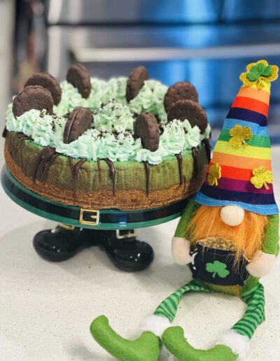 A festive St. Patrick's Day-themed cheesecake topped with mint green whipped cream and chocolate cookies. The cake sits on a stand resembling leprechaun legs. Next to it, there is a gnome figurine wearing a rainbow hat with a four-leaf clover and green-striped pants.