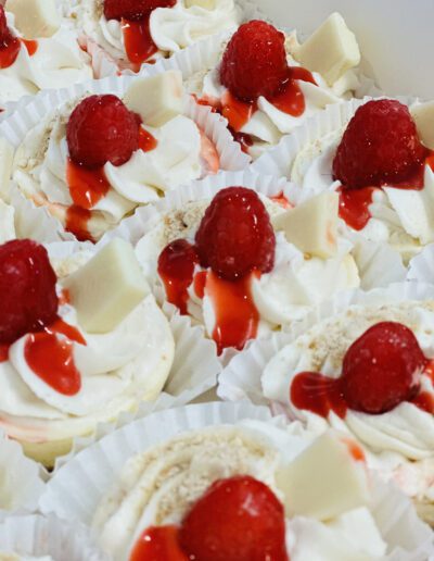 A close-up of multiple white paper-lined mini cheesecakes, each topped with a raspberry, white chocolate pieces, and a drizzle of raspberry sauce. The cheesecakes have a swirled white and pink frosting.