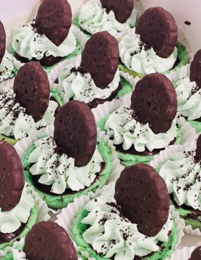 A dozen cupcakes decorated with mint green frosting, topped with chocolate cookies partially embedded in the frosting. The frosting appears swirled and sprinkled with chocolate crumbs. The cupcakes are arranged in white paper liners.