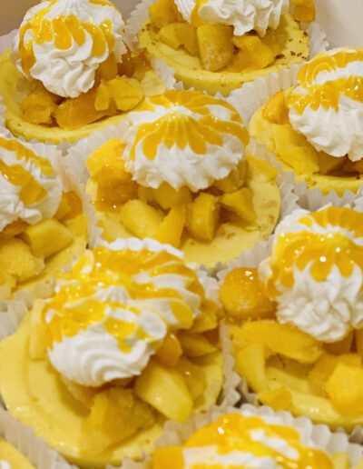 Mini cheesecakes topped with generous dollops of whipped cream, diced mango pieces, and drizzles of mango sauce are arranged closely together. The bright yellow and white colors create a vibrant, appetizing display, ideal for a dessert table or party.