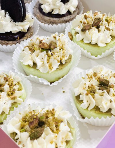 A close-up of a box filled with mini cheesecakes. Some are topped with pistachios and vanilla cream, while others have chocolate cream and Oreo cookies on top. They are placed in white paper cups and arranged neatly in the box.