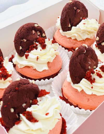 Six pink cupcakes topped with swirls of white frosting, each adorned with a red chocolate cookie and red cake crumbs. The cupcakes are placed in white paper liners and arranged neatly in an open pink box.