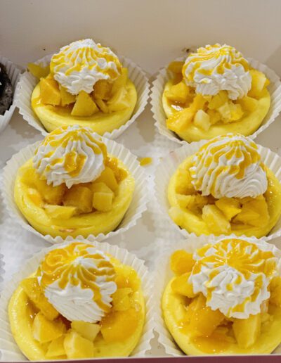 A box containing six mini cheesecakes topped with whipped cream and diced mangoes. The cheesecakes are arranged in white paper cups and drizzled with mango sauce, adding a vibrant yellow color on top.