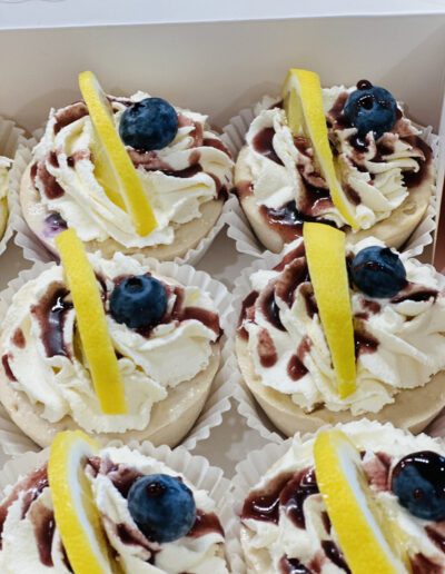 A box of decorative cupcakes topped with generous swirls of white frosting, a drizzle of berry sauce, fresh blueberries, and slices of lemon. Each cupcake is placed in a white cupcake liner and arranged neatly in rows.