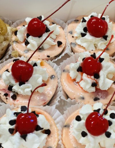 A close-up of six mini cheesecakes topped with whipped cream, chocolate chips, and a cherry. The cheesecakes are in white paper liners and arranged in a box. Their pink-tinted filling suggests a fruity flavor, possibly cherry or strawberry.