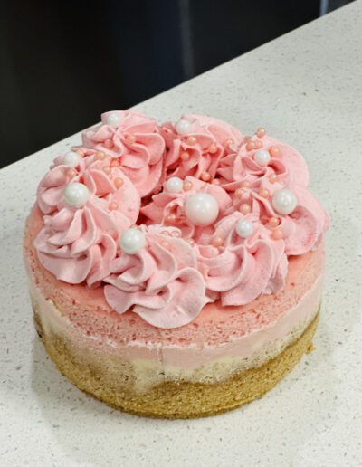 A small, round cake with a layered base of graham cracker crust, cream filling, and pink frosting on top. The cake is adorned with pink and white whipped cream rosettes, and decorated with pearl-like edible beads.