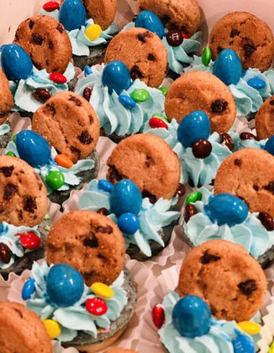 A tray of decorated cupcakes with blue frosting, topped with a chocolate chip cookie, blue candy-coated chocolates, and colorful mini candy pieces. The cupcakes are arranged closely together in white paper liners.