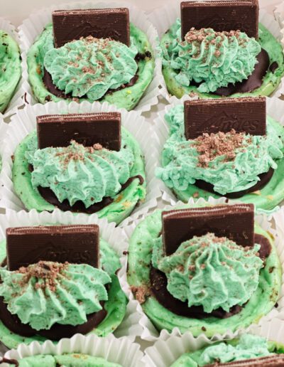 A close-up of eight mint chocolate cupcakes adorned with light green frosting, crumbled mint chocolate bits, and topped with rectangular pieces of dark chocolate. Each cupcake sits in a white paper liner, and the treats are neatly arranged in a box.